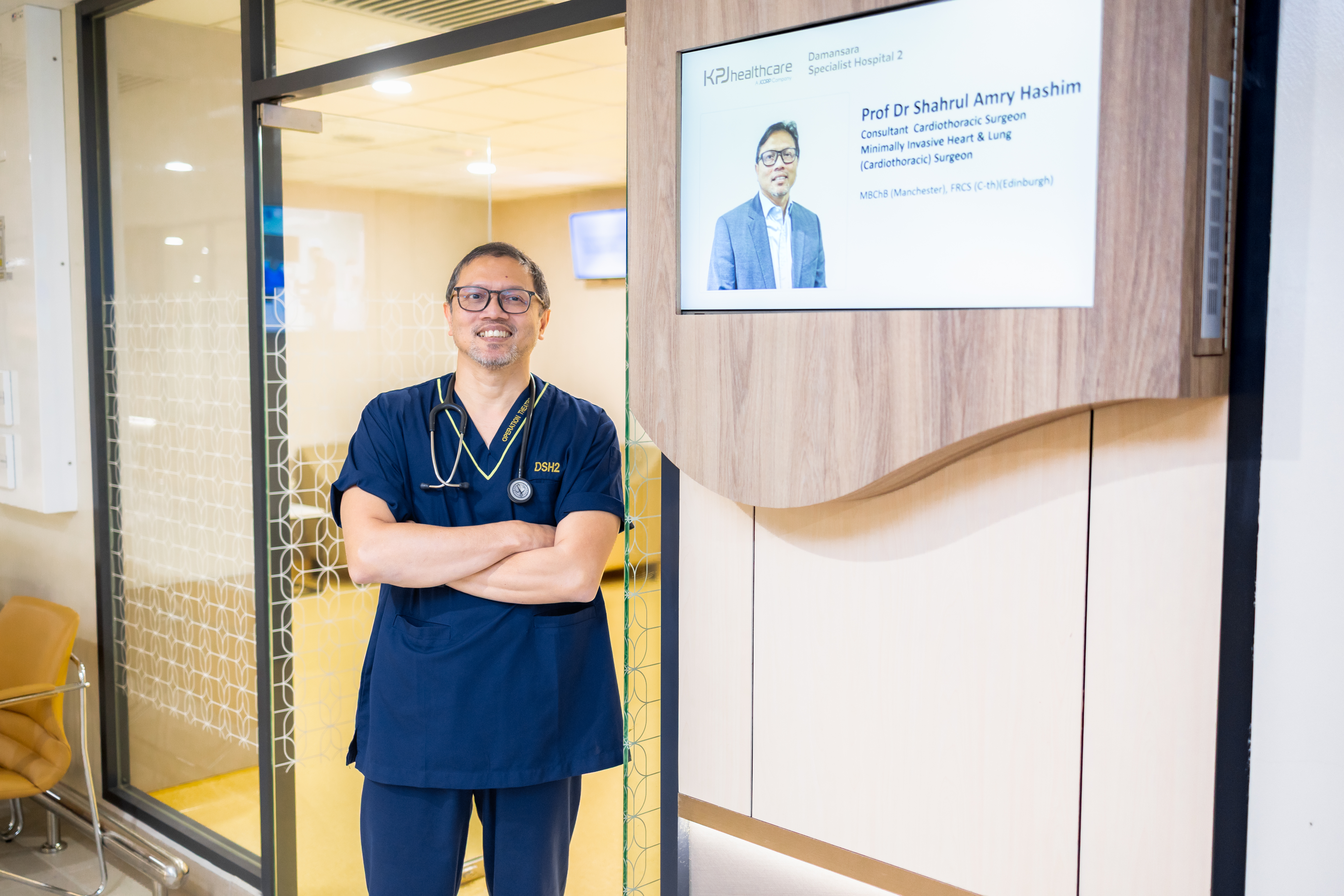 Prof. Dr. Shahrul Amry Hashim standing at the entrance of his clinic at KPJ Damansara Specialist Hospital 2, with his credentials displayed on the screen behind him