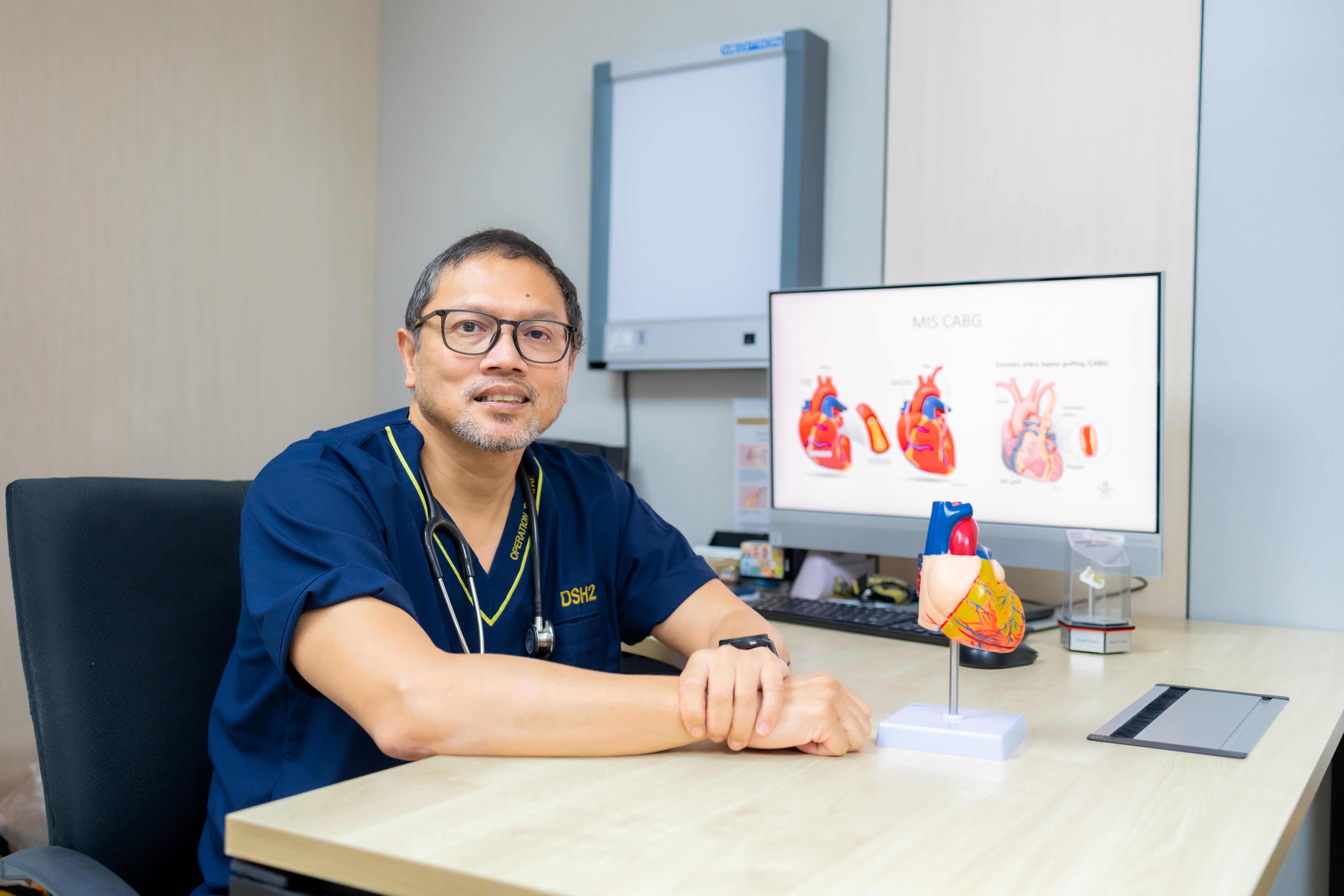 Prof. Dr. Shahrul Amry Hashim, Consultant Cardiothoracic Surgeon at KPJ Damansara Specialist Hospital 2, seated at his desk with a heart model and MIS CABG diagrams