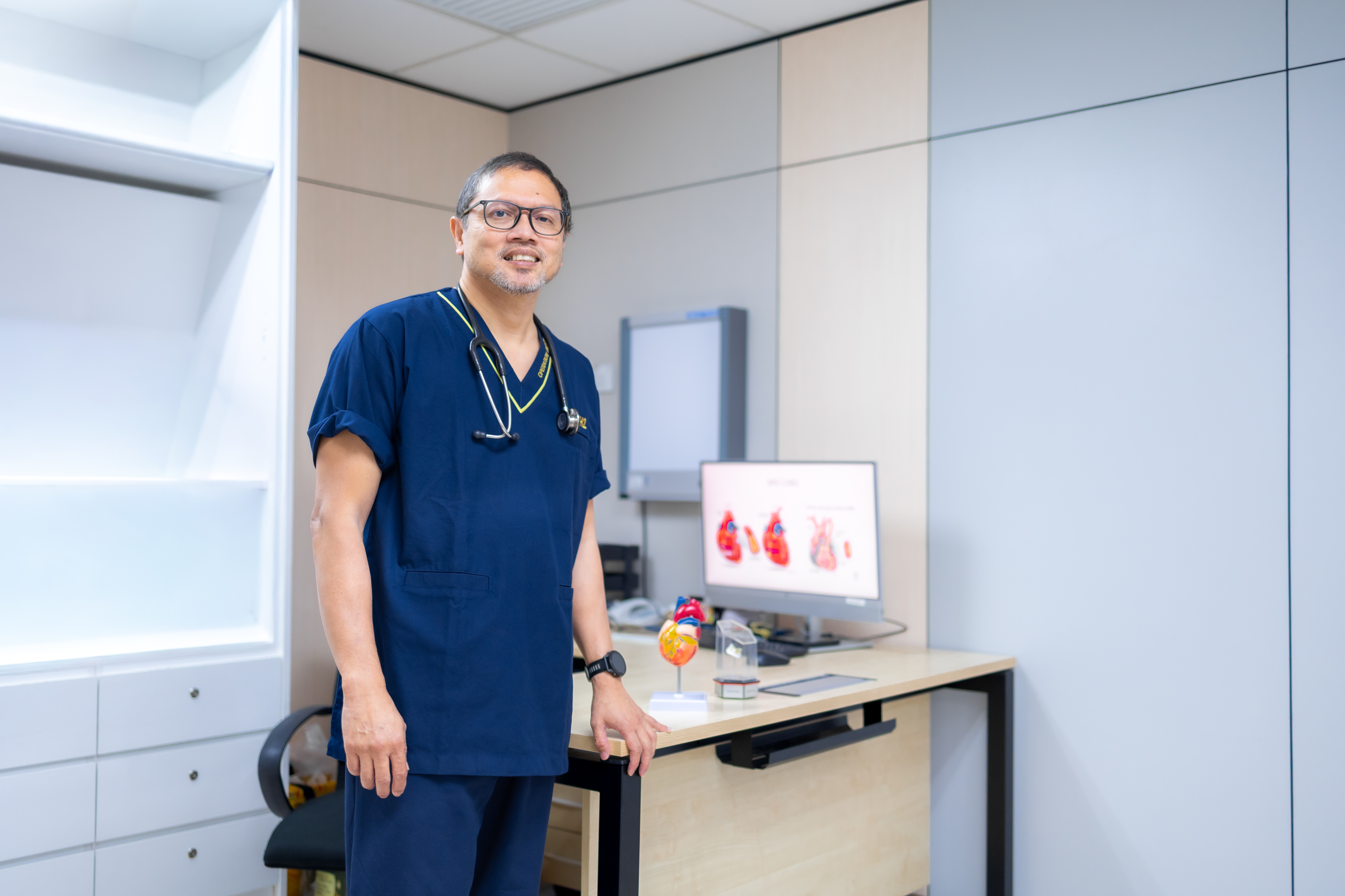 Prof. Dr. Shahrul Amry Hashim standing in his consultation room at KPJ DSH2 with MIS CABG diagrams visible on the monitor behind him
